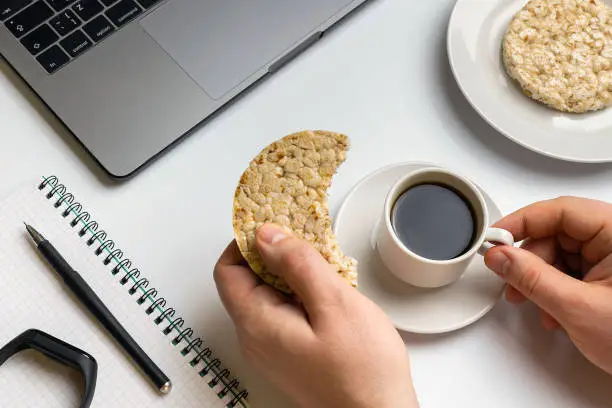 Healthy snacking at work during break time. Sportsman eating crispy rice rounds with peanuts, cup of coffee near the laptop, fitness-tracker and notebook. White organized desk.