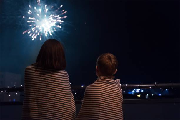 Mother and son looking at New Year celebration fireworks in nigh Mother and son looking at New Year celebration fireworks in night sky warmly wrapped in striped plaids standing on home balcony. new year urban scene horizontal people stock pictures, royalty-free photos & images