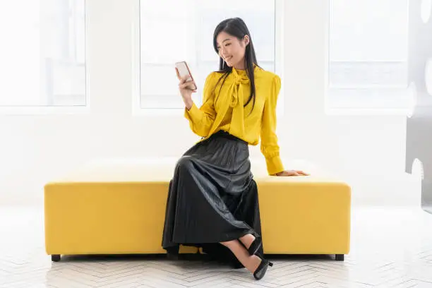 Cheerful mid adult female in smart clothing, resting on mustard colour seat in modern boutique, checking smartphone, retail, shopping, social media