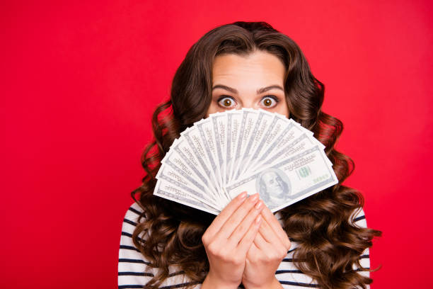 istock Portrait of nice funny attractive frightened wavy-haired lady wearing striped pullover holding in hands hiding behind fan of hundreds usd dollars isolated over bright vivid shine red background 1097372094