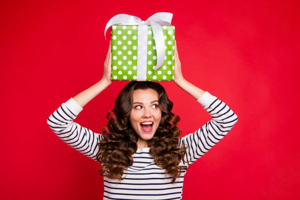 istock Portrait of nice cute winsome attractive cheerful wavy-haired lady wearing striped pullover holding on head large green box opened mouth isolated on bright vivid shine red background 1097374856