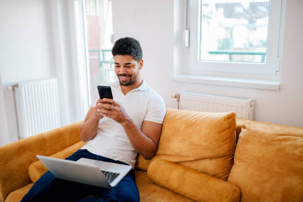 istock Smiling handsome young man texting on mobile phone while sitting on the sofa with laptop in his lap. 1097392966