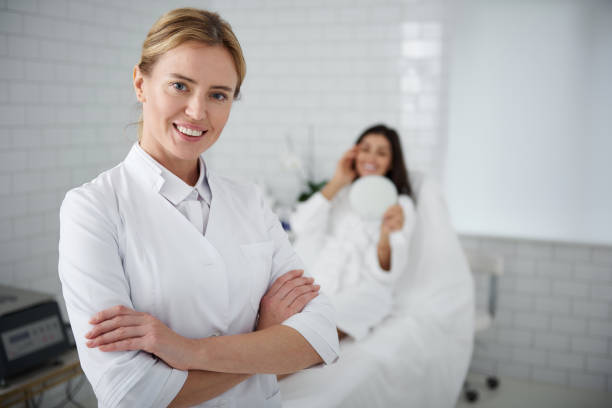 Joyful cosmetologist standing in her cabinet wile client resting on daybed Waist up portrait of charming beautician in white lab coat crossing arms and looking at camera with smile. Young woman in bathrobe looking in the mirror on blurred background beautician stock pictures, royalty-free photos & images