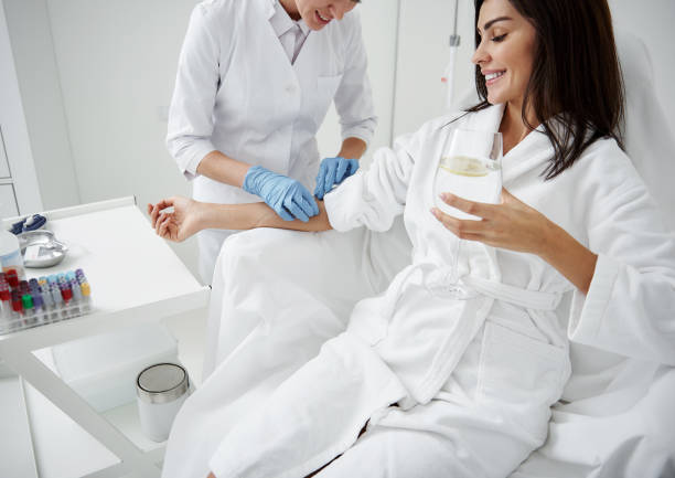 Doctor attaching intravenous drip on lady hand while she drinking water Cropped portrait of beautiful woman in white bathrobe sitting in armchair and receiving IV infusion. She is holding glass of beverage with lemon and smiling human arm stock pictures, royalty-free photos & images