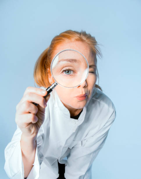 Confident researcher looking through optical Instrument. Female medical professional is using magnifying glass. She is against blue background.