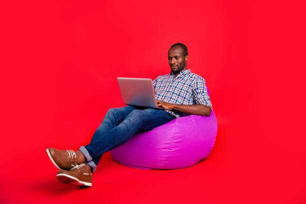 Nice calm positive handsome attractive guy wearing checkered shirt working remotely on laptop sitting on violet bag isolated over bright vivid shine red background