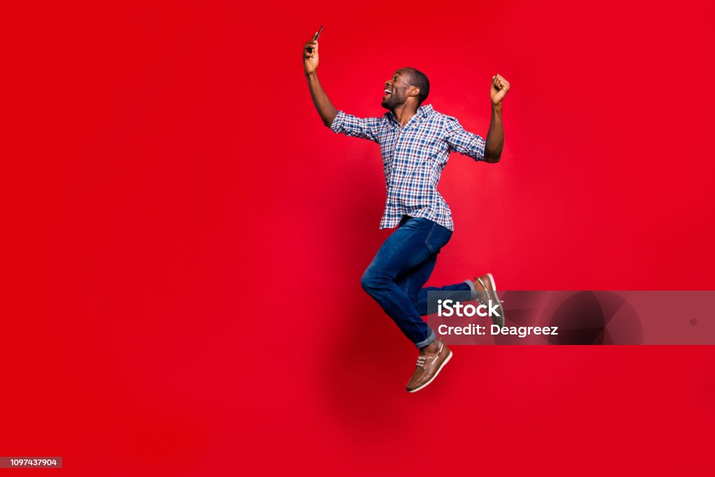 Full length body size profile side view of nice funny crazy handsome cheerful guy wearing checkered shirt holding in hands cell reading text going in air isolated on bright vivid shine red background Jumping Stock Photo
