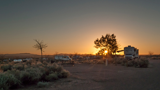 Campground in the desert featuring small trees, cacti and shrubs at sunset. Motor homes and travel trailers are in the distance. Sunset in arid country.