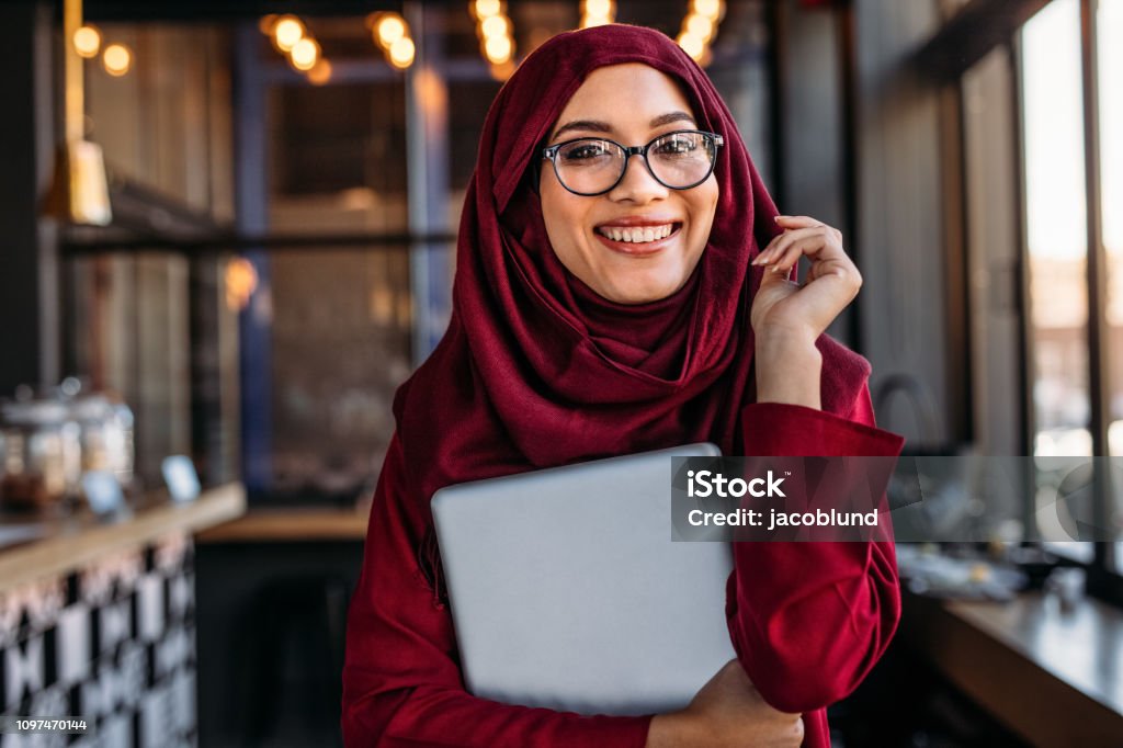 Smiling businesswoman in hijab at coffee shop Pretty young islamic female holding a laptop while at a cafe. Smiling businesswoman in hijab at coffee shop. Islam Stock Photo