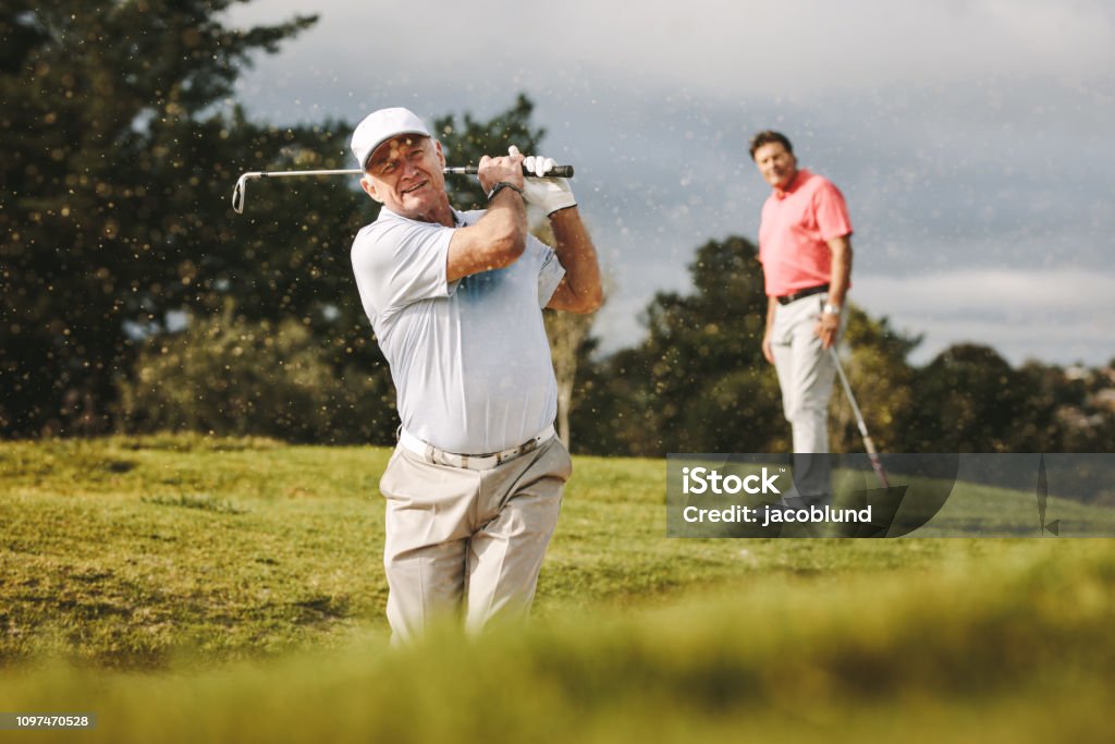 Pro golfer playing on the course Pro golfer playing on the course with second player standing at back looking on. Man hitting the ball out of a sand bunker during the game. Golf Stock Photo