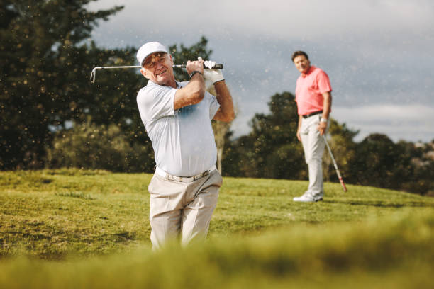 Pro golfer playing on the course Pro golfer playing on the course with second player standing at back looking on. Man hitting the ball out of a sand bunker during the game. senior golfer stock pictures, royalty-free photos & images