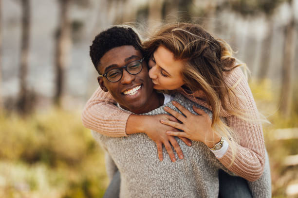 Interracial couple piggybacking outdoors Handsome young man giving piggyback ride to his girlfriend. Couple having fun outdoors. Man carrying his girlfriend on his back, with woman kissing man on the cheek. cheek stock pictures, royalty-free photos & images