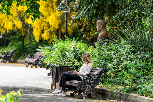 London, UK - June 23, 2018: Stylish one woman people sitting with phone on bench in Chelsea Embankment Gardens by Thames River with green plants and trees