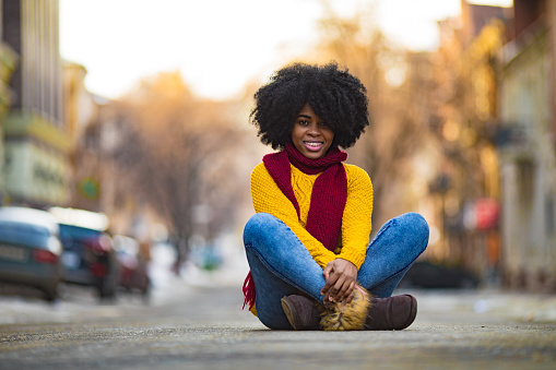 Cheerful young black woman in scarf and sweater is sitting in the middle of the street.