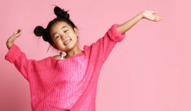 Asian kid girl in pink sweater, white pants and funny buns stands with hands up and smiles. close up stock photo