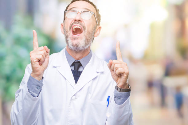 istock Middle age senior hoary professional man wearing white coat over isolated background amazed and surprised looking up and pointing with fingers and raised arms. 1097597708