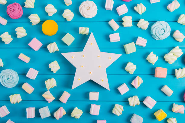 istock White glowing LED star surrounded with colorful square and round marshmallow and macaroons on vibrant blue background. 1097601738