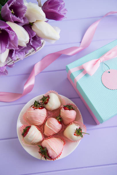 caja de regalo, fresa en chocolate en placa, cinta rosa y bouquet de tulipanes color púrpura y blanco en cesta de mimbre en mesa color lila. - table wedding flower bow fotografías e imágenes de stock