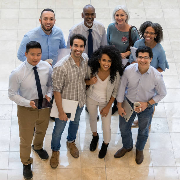 High angle view of cheerful group employee photo A diverse group of coworkers stand together in an office building and look up at the camera and smile.  They are dressed in business casual clothing as they hold paperwork, coffee, a laptop, digital tablet, smart phone and a binder. organised group photo stock pictures, royalty-free photos & images