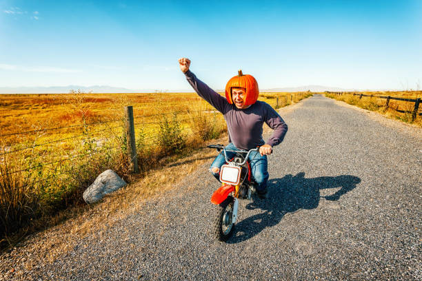 Pumpkin Helmet Motorcycle Racer Photo of an eccentric man wearing a helmet carved from a pumpkin, enthusiastically speeding along on a child-size motorcycle, on a back country road eccentric stock pictures, royalty-free photos & images