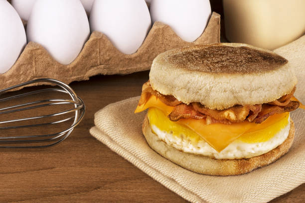 Sandwich breakfast with ingredients in background. English muffin, egg, cheese and bacon.