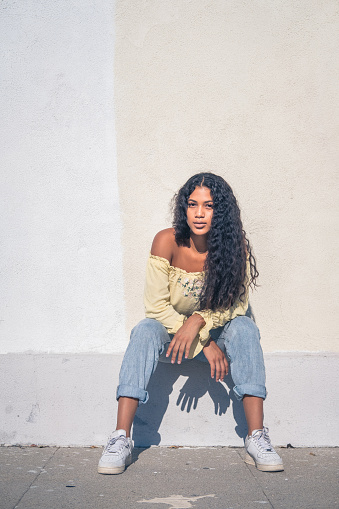 Young millennial female black woman sits on a wall with her elbows leaning on her knees. She is wearing a crop top long sleeve blouse. Her long black curly hair lays on the entire front of her body.
