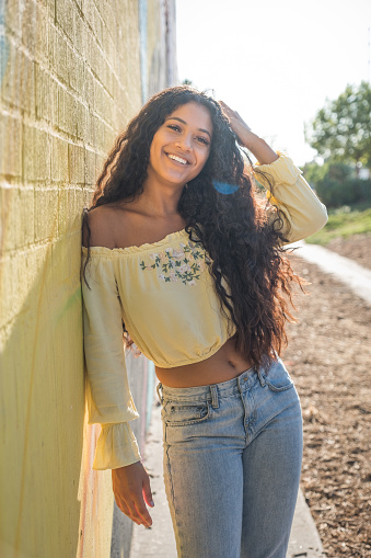 Young millennial black woman stands leaning her side against a yellow wall. She is wearing a crop top long sleeve blouse. Her long black curly hair lays on the entire front of her body. She smiles straight into the camera