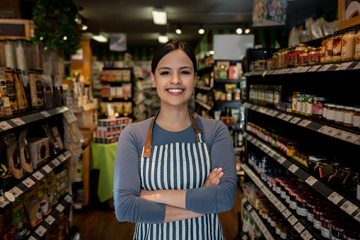Confident female business owner of a supermarket standing between shelves while facing camera smiling with arms crossed