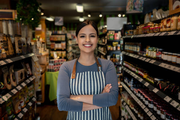 confident female business owner of a supermarket standing between shelves while facing camera smiling - reduction looking at camera finance business imagens e fotografias de stock