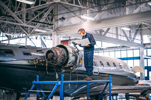 Aircraft mechanic standing on a raised platform, looking at a liquid crystal display of a probe inspection camera, trying to diagnose a fault in a jet engine of a small private airplane in an airport maintenance hangar.