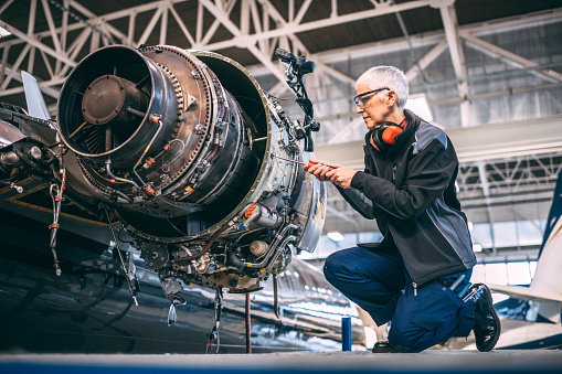 Senior female airplane engineer unscrewing a part of a small jet engine on a private plane during maintenance in an aircraft hangar.