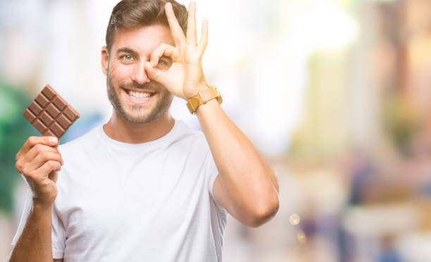 istock Young handsome man eating chocolate bar over isolated background with happy face smiling doing ok sign with hand on eye looking through fingers 1097765792