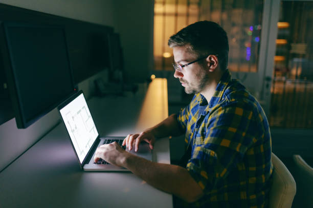 difícil trabalhar sério jovem empregado com óculos e de camisa usando laptop para projeto ficando enquanto sentado no escritório tarde da noite. - esforço - fotografias e filmes do acervo