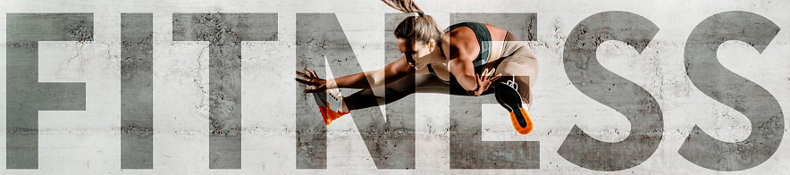 Serious Caucasian woman in sportswear jumping. In background wall. Upper-case fitness over the picture.