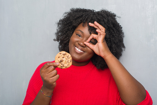 Young african american woman over grey grunge wall eating chocolate chip cooky with happy face smiling doing ok sign with hand on eye looking through fingers