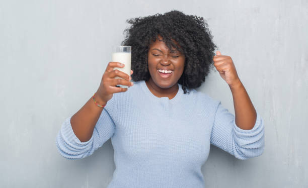 joven mujer afroamericana sobre pared gris grunge beber un vaso de leche gritando orgulloso y celebrar la victoria y el éxito muy emocionado, que anima la emoción - mujer bebiendo leche fotografías e imágenes de stock