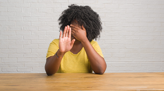 Young african american woman sitting on the table at home covering eyes with hands and doing stop gesture with sad and fear expression. Embarrassed and negative concept.
