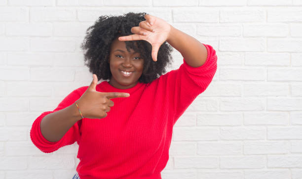 istock Young african american plus size woman over white brick wall smiling making frame with hands and fingers with happy face. Creativity and photography concept. 1097783140