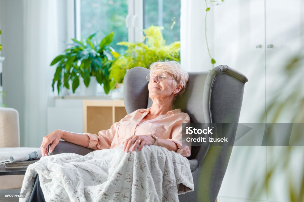 Elderly lady resting in an armchair in her room Pleased senior woman napping in an armchair covered with blanket in her house alone. Senior Adult Stock Photo