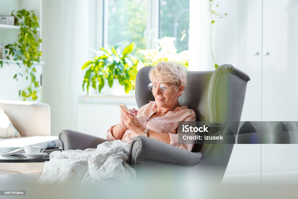 Elderly lady using a smart phone in her room Pleased senior woman sitting in an armchair covered with blanket in her house alone and using a smart phone. Senior Adult Stock Photo