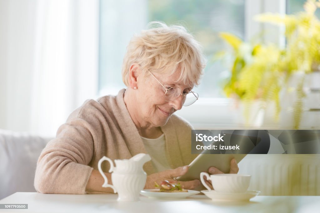 Pleased senior woman using digital tablet Smiling senior woman sitting at the table in her house alone and reading e-book. Senior Adult Stock Photo