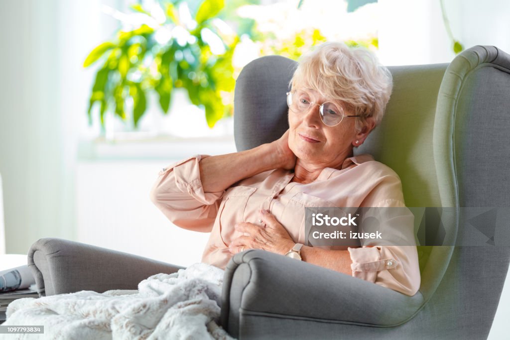 Senior woman having neckache Worried senior woman sitting in an armchair covered with blanket, massaging her neck. Neckache Stock Photo