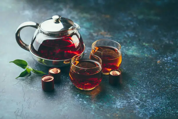 Two glass cups and teapot with black tea and chocolate candies on dark background.