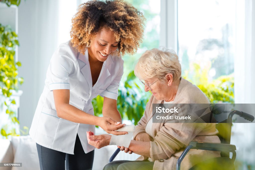 Home nurse bandaging the wrist an elderly lady Afro american female home nurse bandaging the wrist to the senior woman. An elderly lady sitting in a wheelchair. Nurse Stock Photo