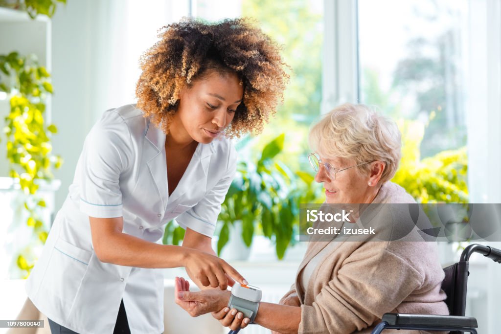 Home nurse checking the blood pressure of the elderly lady Afro American female home nurse measuring the arterial pressure of the senior woman. An elderly lady sitting in wheelchair. Blood Pressure Gauge Stock Photo