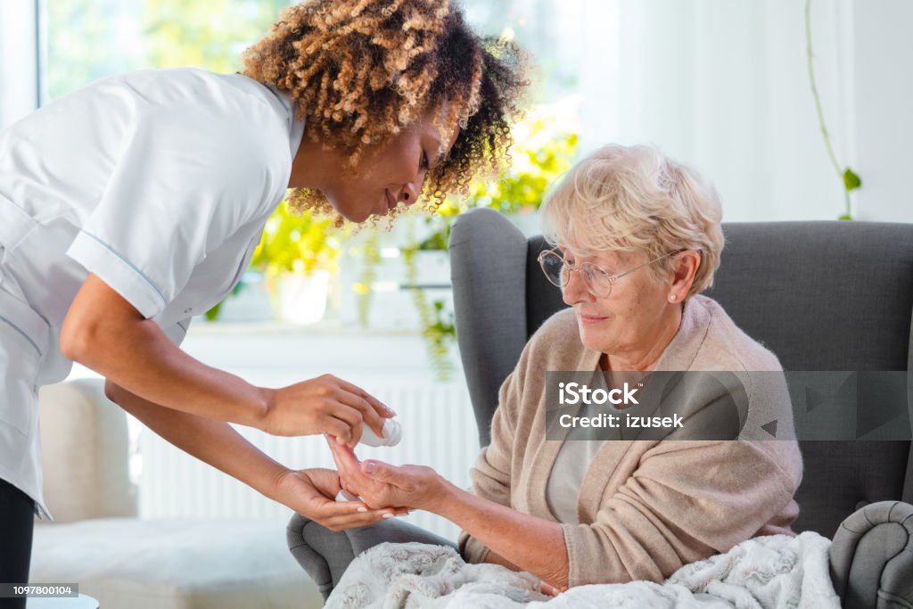 Home nurse giving pills an elderly lady Afro American female home nurse giving medicine to the senior woman. An elderly lady sitting in an armchair. Disability Stock Photo