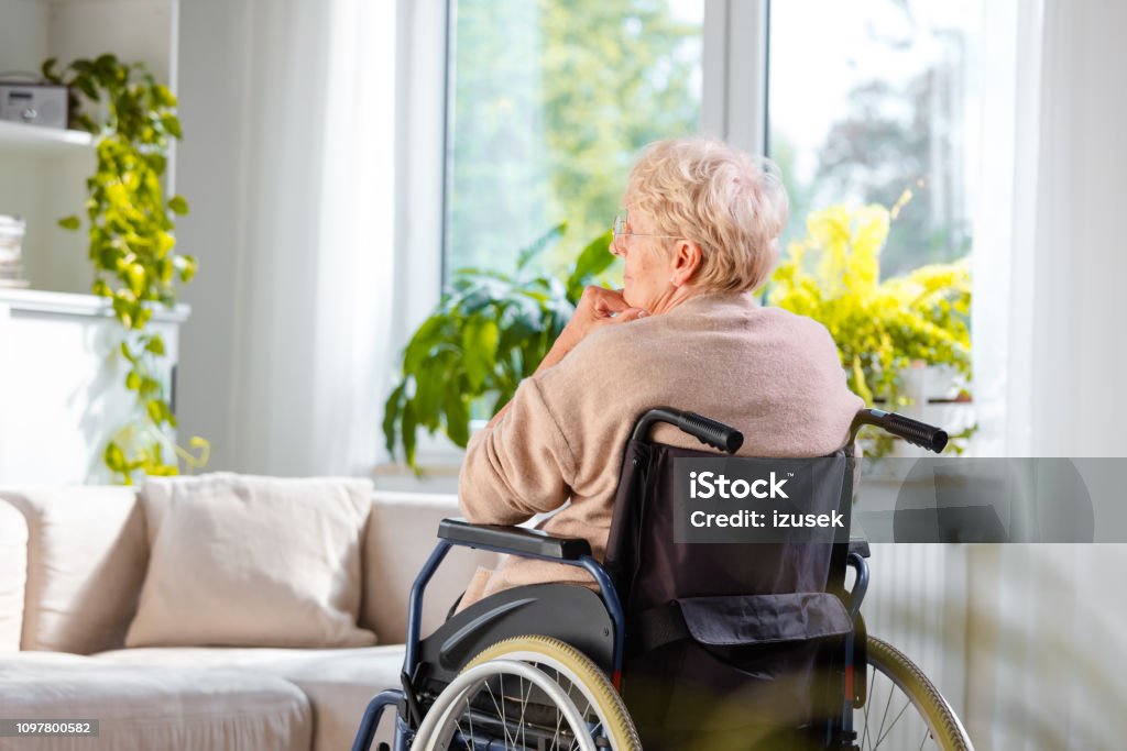 Lonely senior woman sitting in wheelchair in her house Back view of lonely senior woman sitting in a wheelchair in her room. Senior Adult Stock Photo