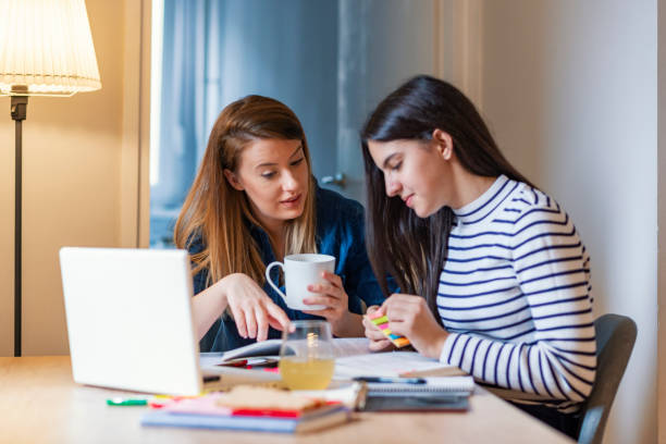 homework time images Involved in her daughter's education Photo of young brown hair Caucasian mother is helping her teen daughter with her homework in front of lap top. They are looking away from the camera. Dark hair schoolgirl and mother together doing homework in home. Beautiful girl and her young mother reading a book together or studying at home during the day. homework table stock pictures, royalty-free photos & images