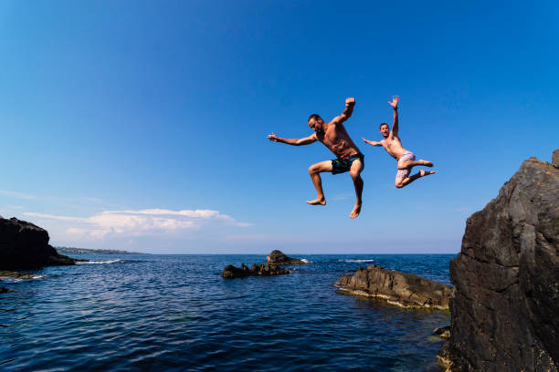 Two young men jumping off cliff into sea Two brave divers jumping off cliff into sea cliff jumping stock pictures, royalty-free photos & images