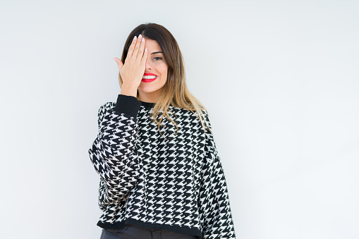 Young woman wearing casual sweater over isolated background covering one eye with hand with confident smile on face and surprise emotion.